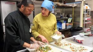 Two chefs in a commercial kitchen are preparing food. The chef on the left, wearing a black coat, is placing ingredients on a tray. The chef on the right, dressed in a yellow coat and a blue headscarf, is holding a container and talking to the other chef.