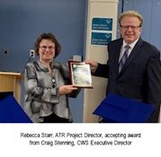 A woman smiles while holding a plaque she received from a man in a suit and tie. They are standing in an office setting with a blue wall and a podium in the background. The caption reads: "Rebecca Starr, ATR Project Director, accepting award from Craig Stenning, CWS Executive Director.