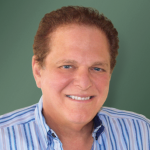 Headshot of Neal Shifman, who has red hair and smiles on a green background. He wears a blue striped shirt.