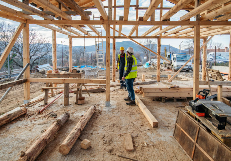 Two construction workers in safety gear and yellow vests are standing inside a wooden house frame under construction. Various building materials and tools are scattered around the site, with a scenic background of trees, hills, and a partly cloudy sky.