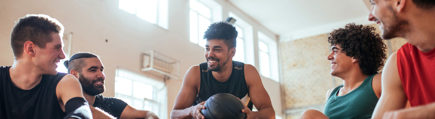 A group of five young men in athletic clothing sit together in a brightly lit gym, smiling and chatting. One of them holds a basketball, indicating they might be taking a break from playing a game. The atmosphere appears relaxed and friendly.