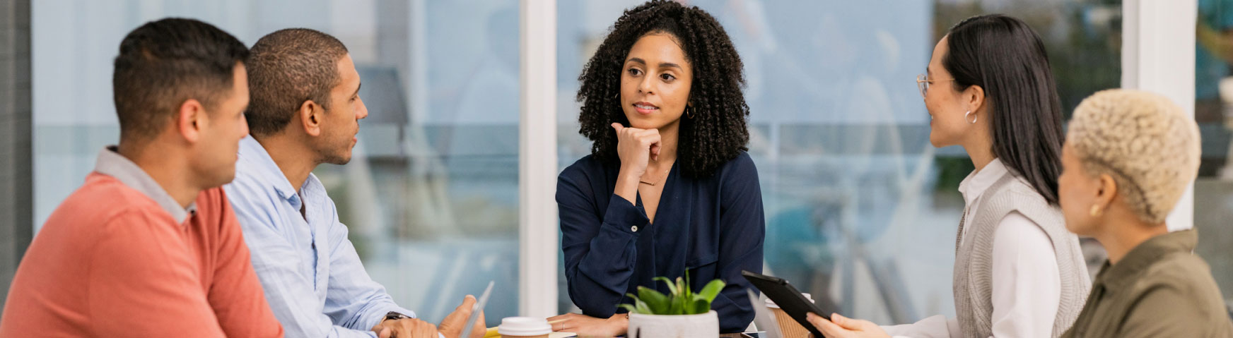 A group of five people are seated around a table engaged in a meeting. One woman, with curly hair and wearing a dark blue top, is speaking while others listen attentively. There is a small potted plant and some drinks on the table. The background is a glass wall.