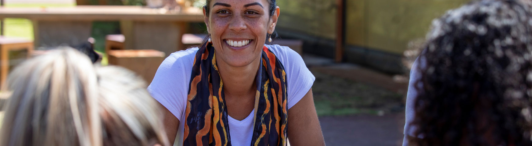 A person in a white shirt and colorful scarf smiles while sitting outdoors. They are engaged in conversation with two people who are partially visible in the foreground. The background features picnic tables and a building.