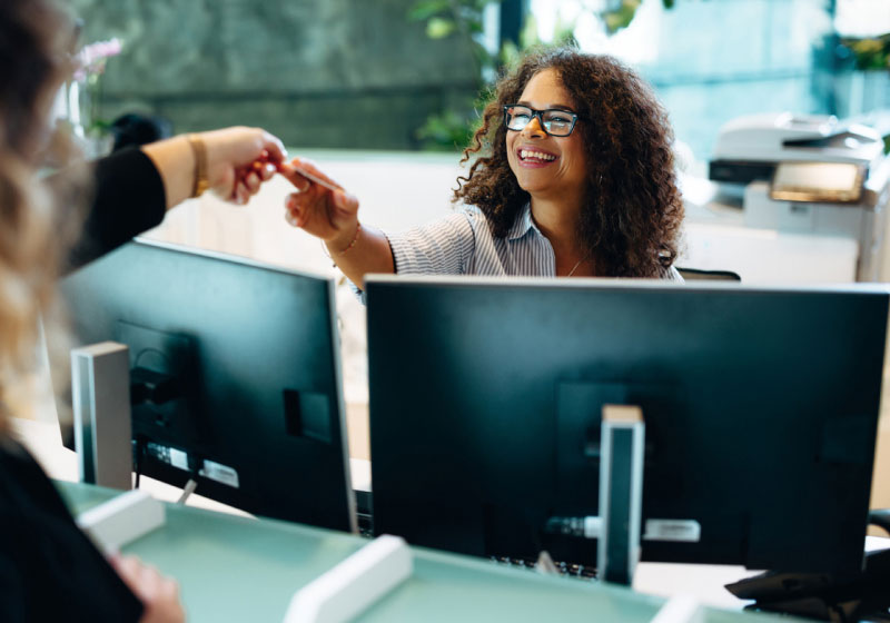 A receptionist with curly hair and glasses, seated at a desk with two monitors, is smiling and handing an item to someone standing off-frame. The background shows office equipment and greenery, suggesting a welcoming office environment.