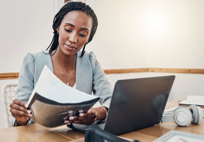 A woman is seated at a desk, reviewing a document in her hands. She has braided hair and is wearing a light gray blazer. In front of her is an open laptop, a set of headphones, and some other office items. The background is a well-lit room with light walls.