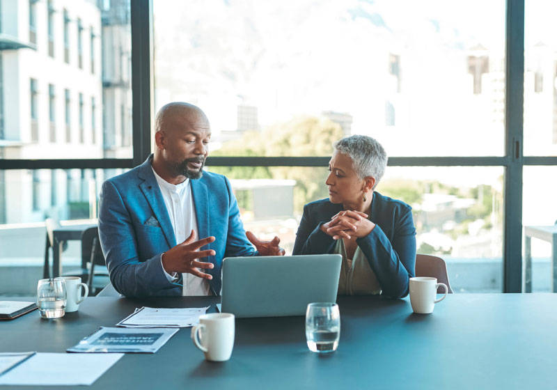 Two business professionals are seated at a conference table, engaged in a discussion. The man on the left is talking with gestures, while the woman on the right listens attentively. A laptop, documents, and glasses of water are on the table. Large windows are in the background.