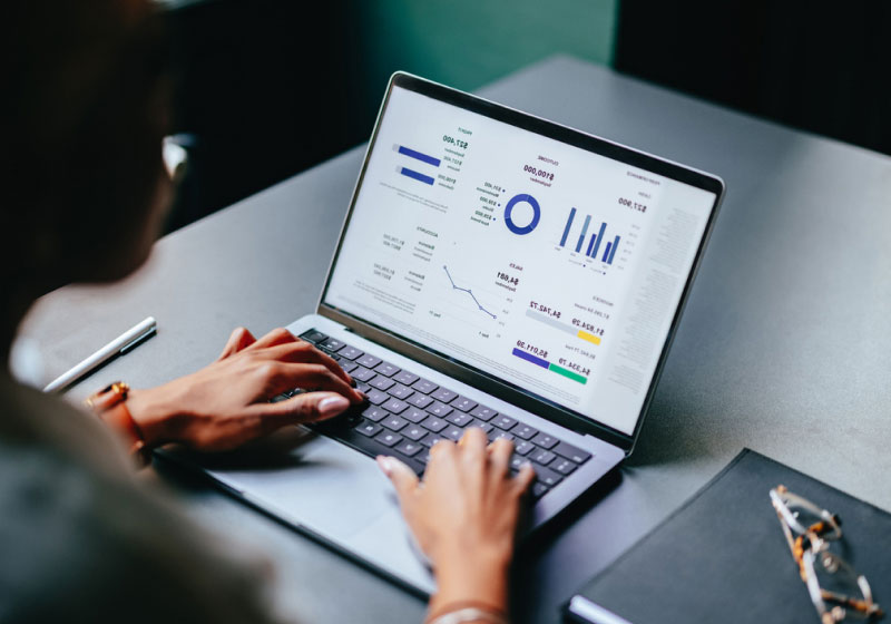 A person is using a laptop displaying various charts and graphs, including bar charts, line graphs, and pie charts with financial data and analytics. The laptop sits on a dark desk next to a closed notebook and a pen.