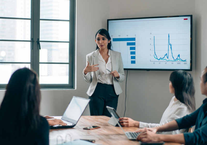 A businesswoman gives a presentation in a conference room with colleagues. She stands in front of a screen showing bar and line graphs, gesturing as she speaks. Her coworkers sit at a table with laptops, attentively listening and taking notes.