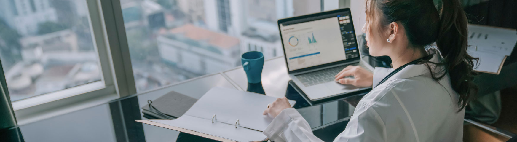 A woman in a lab coat is seated at a desk with a view of a cityscape through a large window. She is working on a laptop displaying charts and graphs, with a binder and a blue coffee mug beside her.