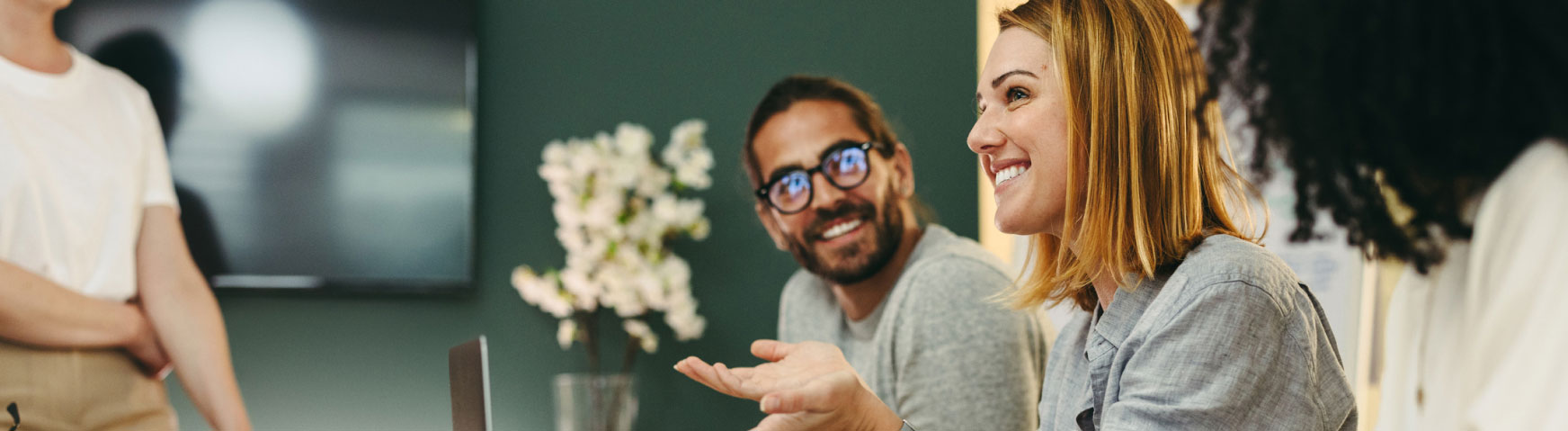 A woman with shoulder-length hair is smiling and gesturing with her hand during a meeting. A man next to her, wearing glasses, is smiling at her. They are at a table with laptops and a vase of flowers in the background.