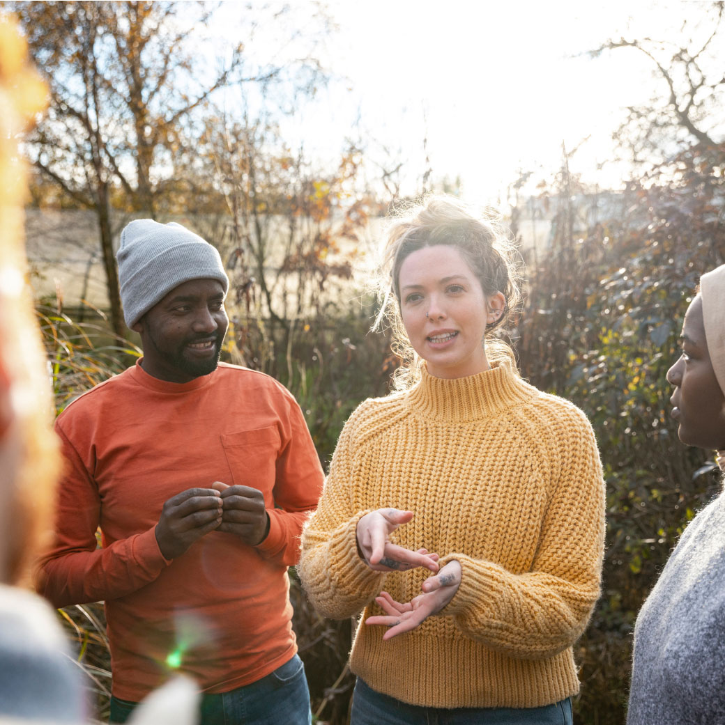 A group of people stands outdoors in a forest, smiling and engaged in conversation. One person in a yellow sweater gestures while speaking, and another in an orange shirt and gray beanie listens attentively. Sunlight filters through trees in the background.