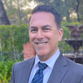 The image shows a headshot of Mark Faucette with short dark hair, smiling. They are wearing a gray suit with light blue shirt and dark blue tie. The background is outside with a fountain and green vines.