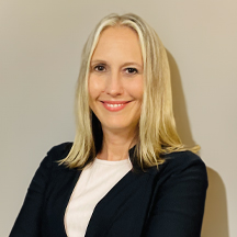 Sabrina Nansteel-Bunt, a woman with long blonde hair, wearing a black blazer and a white top, is standing against a plain light-colored wall. She is smiling and has her arms crossed.