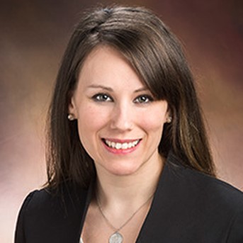 Jennifer is pictured with straight brown hair, smiling, wearing a black blazer over a white top, and a necklace. She has fair skin, pearl earrings, and is posed against a warm, blurred background.