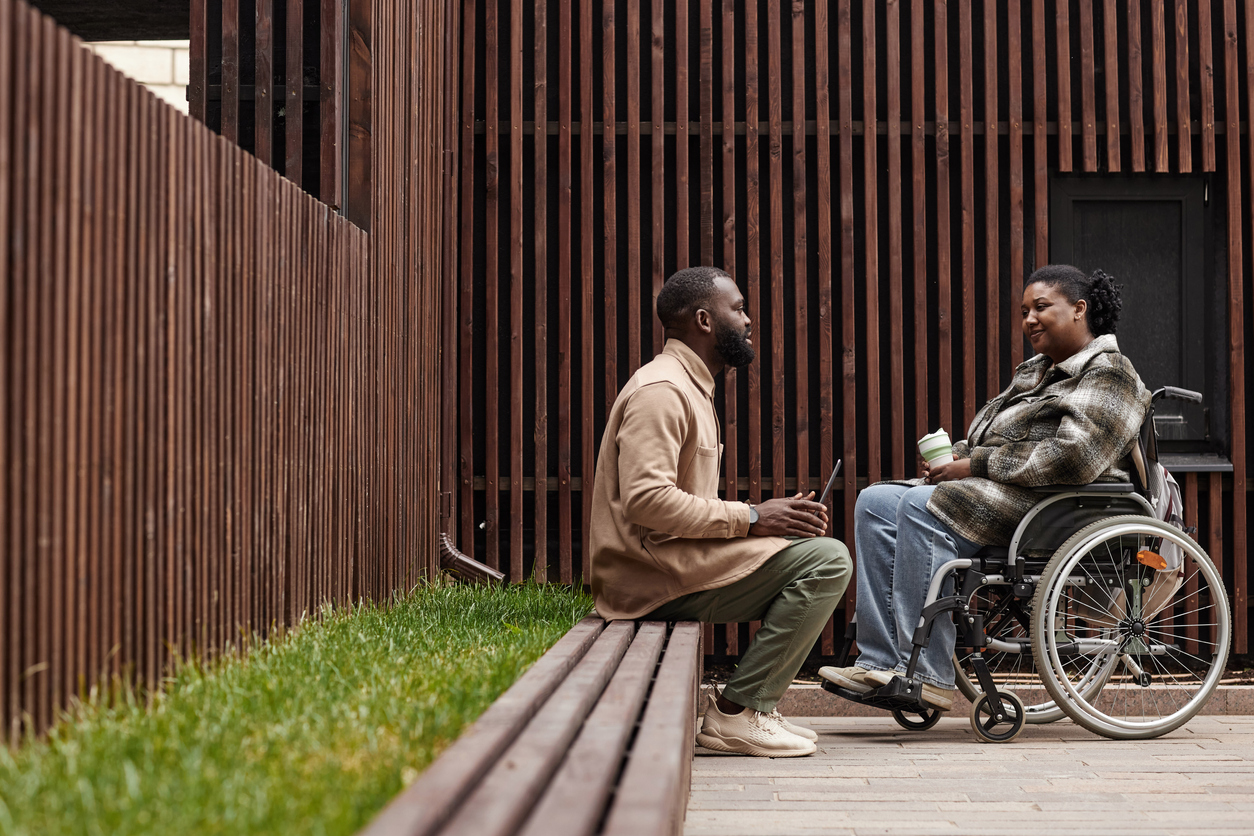 A man sits on a bench having a conversation with a woman in a wheelchair outdoors. They are near a wooden fence and building, with grass beside the bench. The woman holds a coffee cup, and both seem engaged in the dialogue.