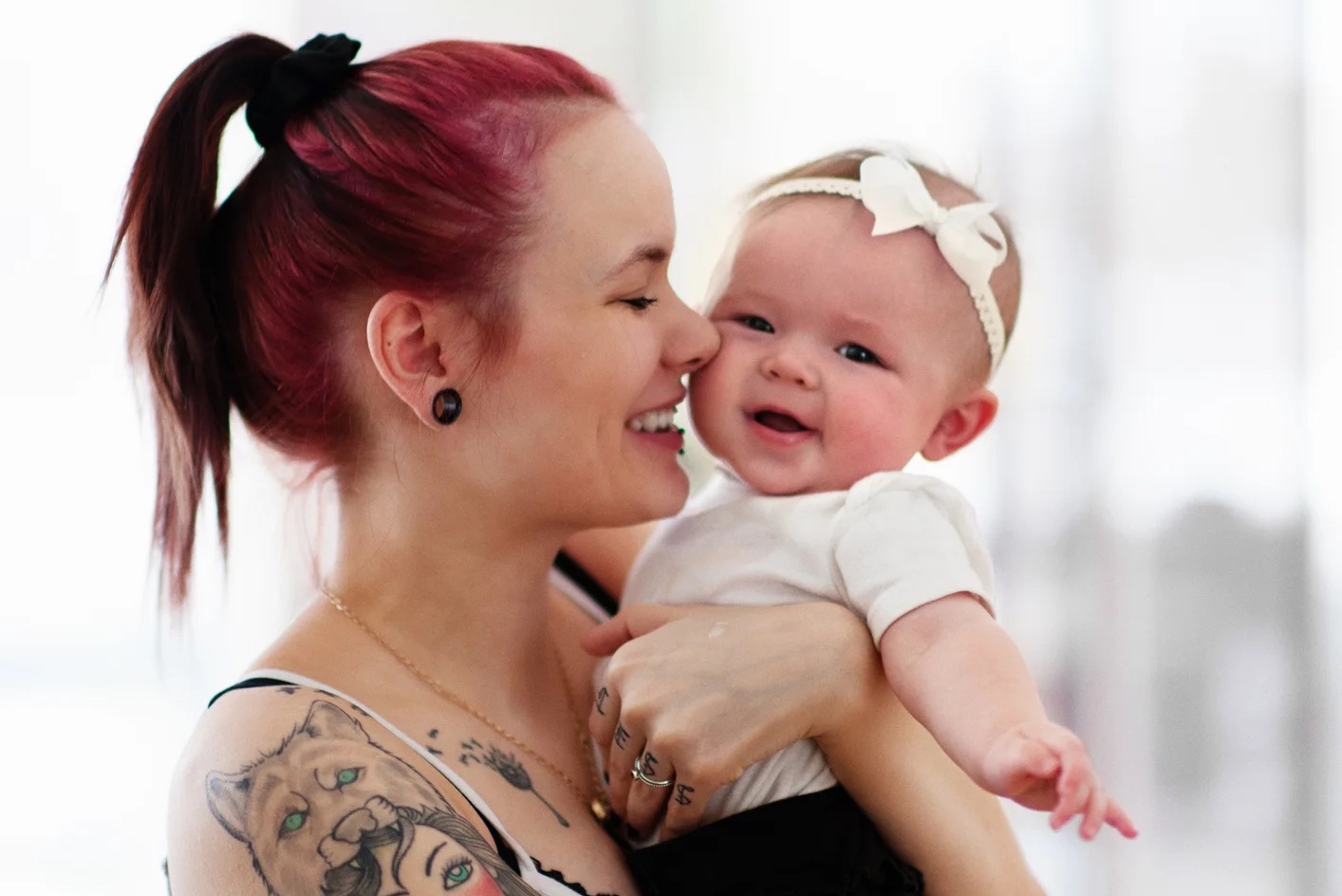 A woman with red hair tied back in a black scrunchie, tattoos on her arms, and wearing a sleeveless top holds a smiling baby girl dressed in a white outfit with a white bow headband. They are both facing each other and smiling, sharing a joyful moment.