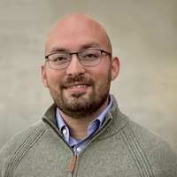 Brent Kaplan, a man wearing wire-rimmed glasses and a green sweater smiles at the camera in front of a soft green background.